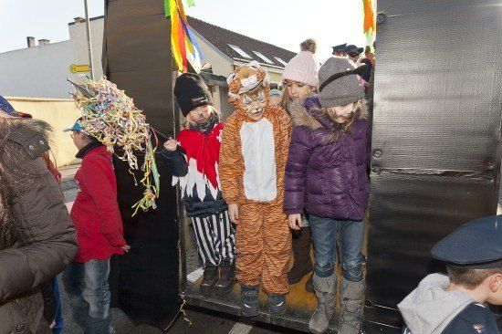 Vier Kinder in Kostümen, darunter ein Tiger, stehen in einem dekorierten Fahrzeug, während andere zusehen.