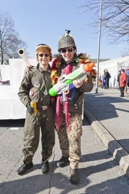Zwei Männer in Militäruniformen und mit Wasserpistolen, die auf einer Straße stehen, mit einem verschwommenen Hintergrund von Menschen und einem Gebäude.