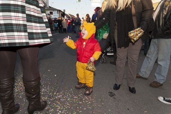 Ein Kind, das als Winnie Puuh verkleidet ist, steht während einer Parade auf der Straße, hält eine Tüte mit Süßigkeiten und ist von Konfetti umgeben.