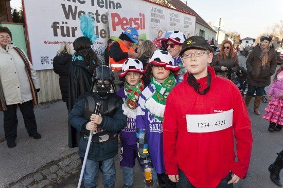 Eine Gruppe von Kindern und Erwachsenen in verschiedenen Kostümen steht auf einer Straße, einige tragen Brillen und Hüte. Ein Banner mit deutschem Text ist im Hintergrund sichtbar.