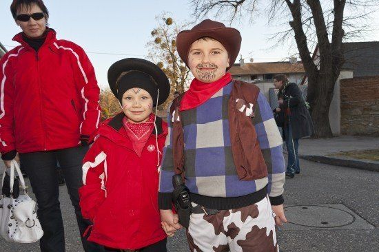 Zwei Kinder in Cowboy-Kostümen, eines mit Gesichtsbemalung, stehen zusammen mit einer erwachsenen Frau in einer roten Jacke auf einer Straße.