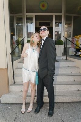 Ein Mann und eine Frau stehen auf den Stufen eines Gebäudes, beide formell gekleidet, der Mann mit Sonnenbrille und die Frau in einem weißen Kleid.