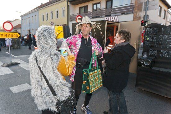 Drei Personen stehen auf einer Straße, einer trägt einen Strohhut, ein anderer ein pelziges Kostüm und der Dritte einen schwarzen Mantel. Sie unterhalten sich, während eine Menschenmenge und Gebäude im Hintergrund sind.