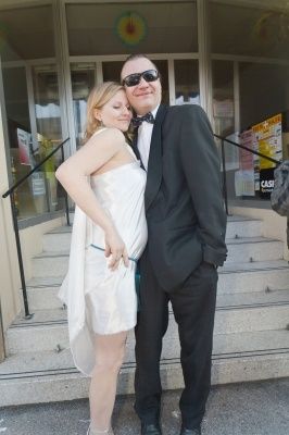 Ein Mann und eine Frau, beide formell gekleidet, stehen auf den Stufen eines Gebäudes. Der Mann trägt eine Sonnenbrille und eine Fliege, die Frau ein weißes Kleid.