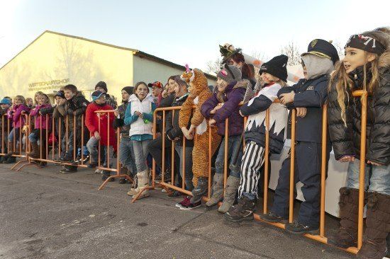 Eine Gruppe von Kindern in Kostümen steht hinter einem Barriere und beobachtet eine Veranstaltung oder einen Umzug. Einige tragen Hüte, Stiefel und Masken.