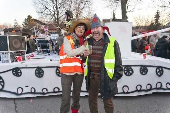 Ein Mann und eine Frau in Sicherheitswesten und Hüten stehen vor einem dekorierten Wagen mit einem Schlagzeug-Set und einer Menschenmenge im Hintergrund.