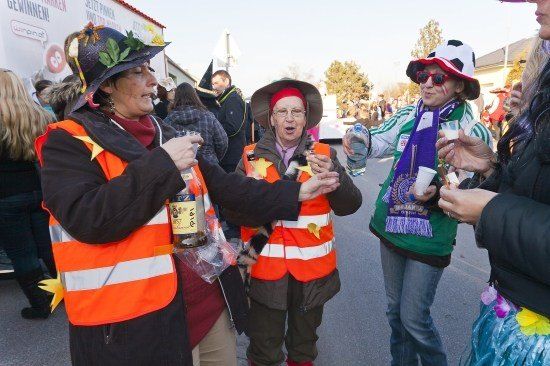 Eine Gruppe von Menschen in festlicher Kleidung, einschließlich Sicherheitswesten und Hüten, versammelt sich auf einer Straße, möglicherweise für eine Parade oder Feier. Sie halten Getränke in den Händen und scheinen sich zu amüsieren.