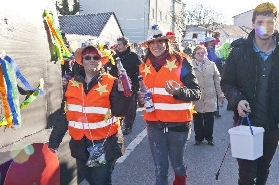 Zwei Frauen in orangenen Westen mit Sternen gehen lächelnd die Straße entlang und halten Flaschen. Ein Mann mit einem Eimer folgt ihnen.