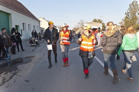 Eine Gruppe von Menschen in Kostümen und hohen Stiefeln geht eine Straße entlang, einige halten Flaschen und andere tragen Taschen. Im Hintergrund ist ein Gebäude mit einer grünen Tür und einem Kinderwagen zu sehen.