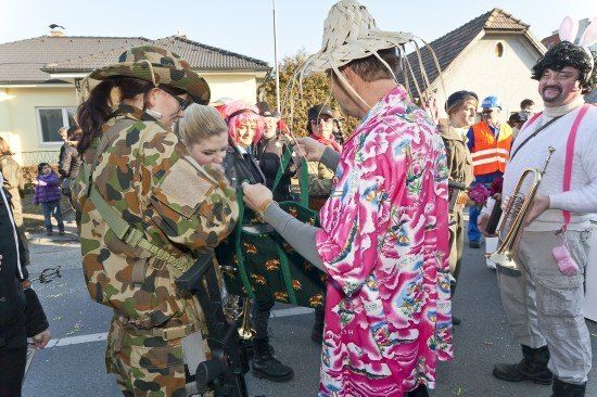 Eine Gruppe von Menschen in verschiedenen Kostümen, darunter eine Camouflage-Uniform und ein rosa Blumenmantel, stehen in einem Außenbereich. Eine Person hält eine Trompete.
