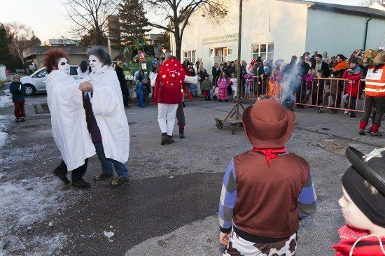 Eine Gruppe von Menschen in Kostümen, darunter zwei in weißen Roben, tanzt vor einer Menschenmenge während einer Winterveranstaltung. Ein Kind in einem Cowboyhut beobachtet.