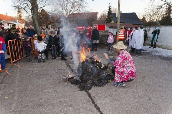 Eine Menschenmenge, einige in Kostümen, versammelt sich um ein Lagerfeuer im Freien. Eine Person in einem rosa Kleid kniet neben dem Feuer.