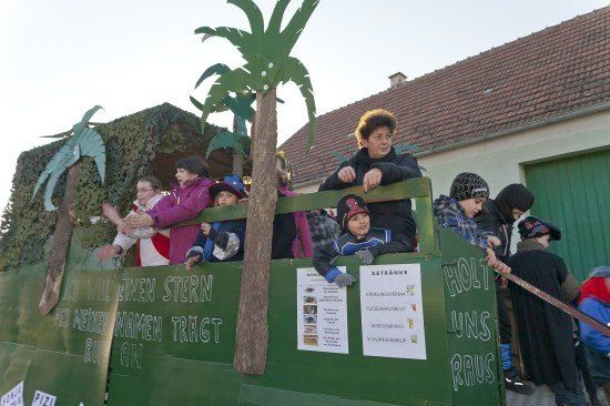 Eine Gruppe von Kindern und Erwachsenen fährt auf einem geschmückten Wagen, möglicherweise für einen Umzug. Der Wagen hat ein tropisches Thema mit einer Palme und einem Dinosaurier. Die Kinder tragen Winterkleidung und halten sich an den Geländern fest. Der Wagen hat ein Schild mit den Worten 'Holy Uns Raus' darauf.