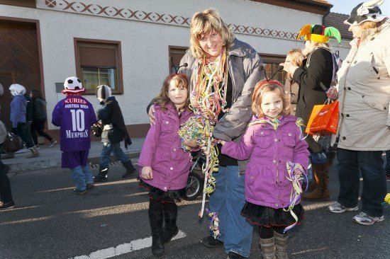 Eine Frau und zwei junge Mädchen in lila Jacken stehen auf einer Straße während eines Umzugs. Sie halten bunte Bänder. Hinter ihnen laufen mehrere Leute, und eine Person trägt einen Hut mit einem Fußball darauf.