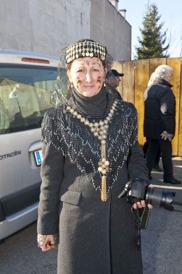 Eine Frau in einem langen grauen Mantel mit einer Perlenkette und einem Hut steht vor einem Van und hält eine Kamera. Hinter ihr ist ein Holzzaun und eine Person in einem schwarzen Mantel zu sehen.