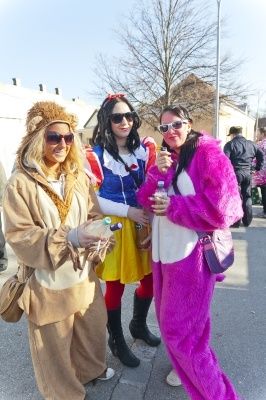 Drei Frauen in Kostümen stehen auf einer Straße, eine verkleidet als Löwe, eine andere als Schneewittchen und die dritte in einem rosa Pelzoutfit. Sie tragen alle Sonnenbrillen und halten Flaschen.