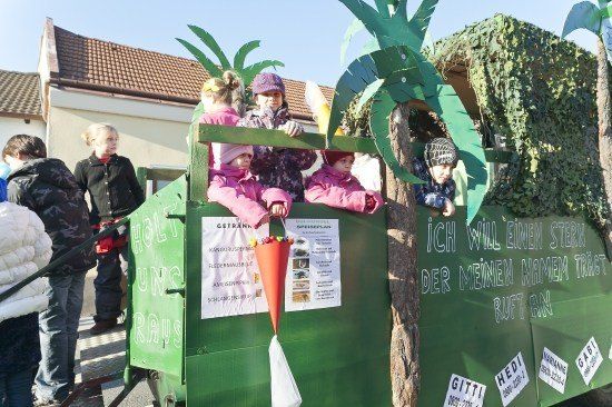 Kinder in rosa Jacken sitzen in einem dekorierten grünen Wagen mit Palmblättern und einem Banner, das 'Ich will einen Stern der meinen Namen trägt' besagt. Eine Person hält einen roten Trichter mit Süßigkeiten.