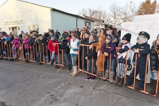 Eine Gruppe von Kindern und Erwachsenen in Kostümen steht hinter orangefarbenen Barrieren und beobachtet eine Veranstaltung vor einem Gebäude mit einem Schild, das 'Wirtschaftshof' besagt.
