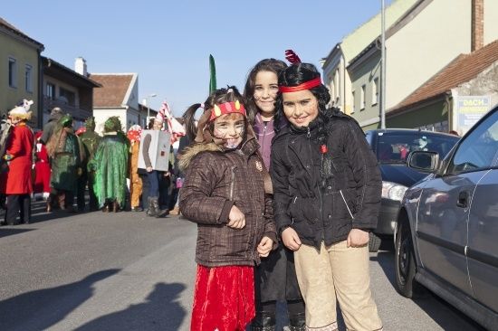 Drei Kinder in Kostümen stehen auf einer Straße, mit einem Umzug im Hintergrund. Die Kinder lächeln und posieren für ein Foto.