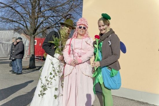 Drei Menschen in Kostümen stehen zusammen draußen, einer in einem weißen Kleid, einer in einem rosa Kleid und einer in einem grünen Hut. Sie lächeln und posieren für ein Foto.