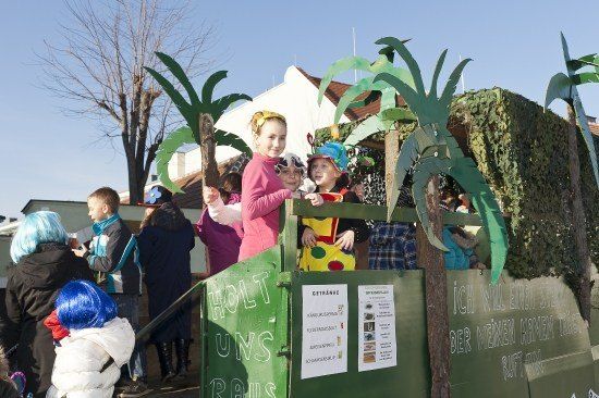Eine Gruppe von Kindern in Kostümen steht auf einem Wagen mit Palmen und einem Schild, das 'Holt Uns' sagt. Sie sind von anderen Menschen umgeben.