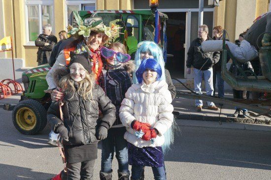 Eine Gruppe von Kindern und einer erwachsenen Frau in Kostümen stehen vor einem Gebäude und einem Traktor. Die Kinder tragen Gesichtsbemalung und Perücken.
