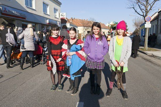 Vier Mädchen in Kostümen stehen auf einer Straße mit vorbeigehenden Menschen und Gebäuden im Hintergrund.