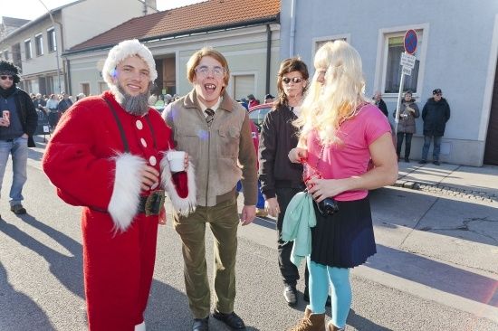 Eine Gruppe von Menschen in Kostümen, darunter ein Mann in einer Santa-Claus-Verkleidung, geht auf einer Straße. Hinter ihnen hält eine Frau in einem rosa T-Shirt eine Kamera. Im Hintergrund befinden sich Gebäude und eine Menschenmenge.