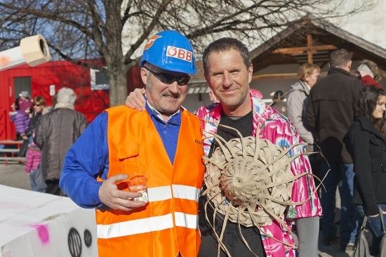 Zwei Männer stehen zusammen in einem öffentlichen Bereich, einer trägt eine reflektierende orange Weste und einen blauen Schutzhelm, der andere hält ein großes dekoratives Objekt. Hinter ihnen steht eine Gruppe von Menschen in der Nähe eines roten Gebäudes und eines Baumes.
