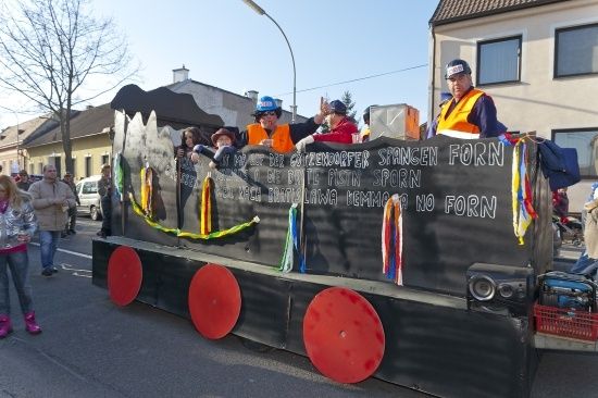 Ein geschmückter Wagen mit Menschen in orange Westen und Hüten fährt während eines Umzugs eine Straße hinunter. Der Wagen hat einen schwarzen Hintergrund mit weißem Text und roten Rädern.