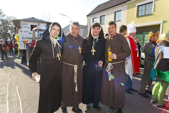 Vier Männer, die als Mönche gekleidet sind, stehen auf einer Straße zusammen, einer hält einen gelben Becher. Dahinter ein Gebäude mit einem Banner und Menschen in verschiedenen Kostümen.