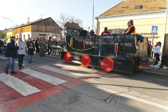 Ein Paradewagen mit Menschen darauf, dekoriert mit Bannern und roten Rädern, bewegt sich auf einer Straße, an der eine Menschenmenge zuschaut.
