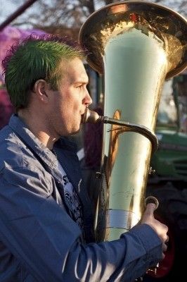 Ein Mann mit grünen Haaren spielt eine große Messingtuba im Freien, wobei ein Traktor im Hintergrund zu sehen ist.