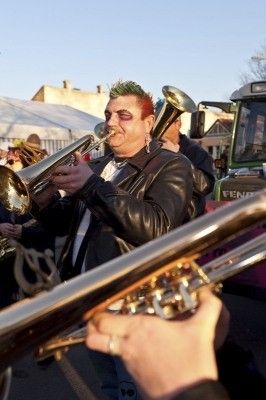 Ein Mann mit grünen Haaren und Gesichtsbemalung spielt eine Posaune bei einer Straßenaufführung.