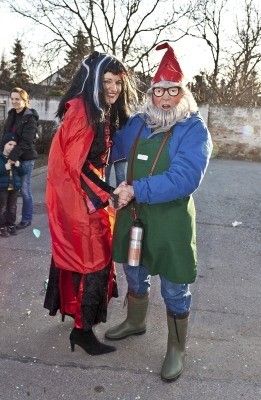 Zwei Menschen in Kostümen schütteln sich die Hände in einer Außenumgebung. Die Frau trägt ein rotes und schwarzes Kostüm mit Hut, und der Mann trägt ein blaues und grünes Kostüm mit Brille und Hut. Hinter ihnen stehen einige Menschen, und einige Bäume sind im Hintergrund.