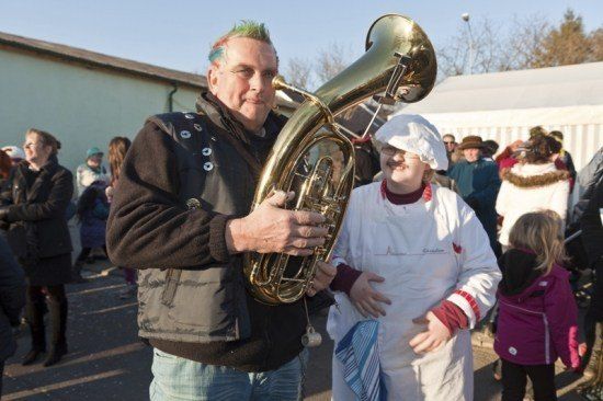 Ein Mann mit grünen Haaren und einem Tuba steht mit einer Frau in einer weißen Schürze bei einer Veranstaltung im Freien. Dahinter steht eine Menschenmenge und schaut zu.