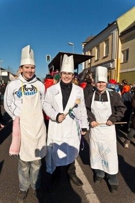 Drei Personen in Kochmützen und Schürzen, stehen auf einer Straße mit Gebäuden und einer Menschenmenge im Hintergrund.