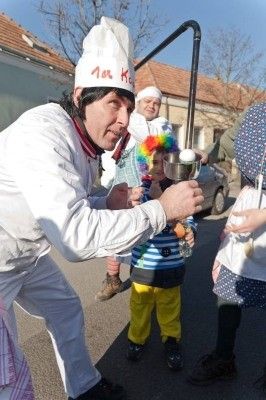 Ein Koch in weißer Uniform und Hut hält eine Schüssel und scheint ein Ei für ein Kind in einem bunten Hut zuzubereiten.