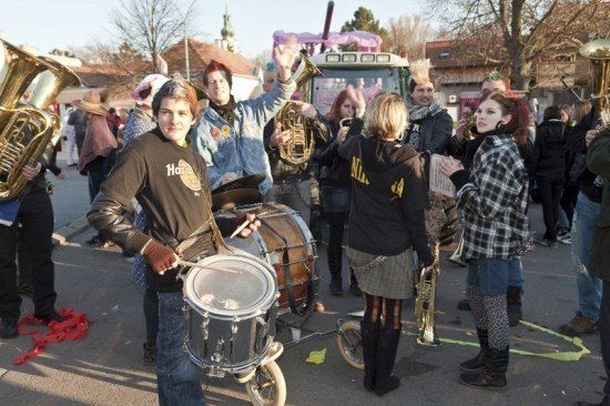 Eine Gruppe von Menschen in verschiedenen Kostümen spielt Musikinstrumente auf der Straße. Ein Mann spielt die Trommeln, während andere Trompeten, Tubas und andere Instrumente spielen. Ein Traktor ist hinter ihnen geparkt.