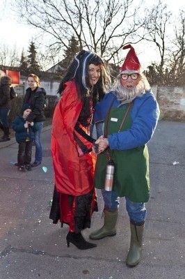 Zwei Menschen in Kostümen, einer in Rot und der andere in Blau und Grün, schütteln sich die Hände im Freien. Hinter ihnen stehen einige Leute, und ein Kind hält ein blaues Objekt. Bäume und ein Gebäude sind im Hintergrund.