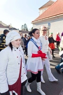 Drei Frauen in weißen Kostümen mit roten Akzenten gehen während eines Umzugs eine Straße entlang, mit Menschen und Gebäuden im Hintergrund.