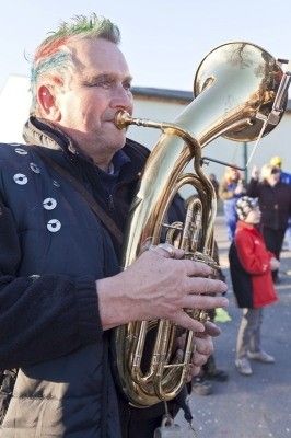 Ein Mann mit grünen Haaren und einem schwarzen Jackett spielt eine Tuba im Freien, mit mehreren Personen im Hintergrund.