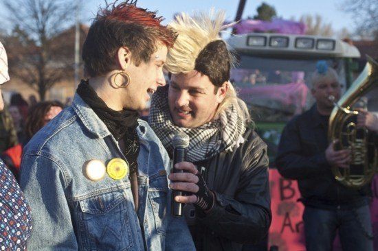 Zwei Männer unterhalten sich mit einem Mikrofon bei einer öffentlichen Veranstaltung, einer trägt eine Jeansjacke und der andere eine schwarze Jacke mit Schal. Dahinter spielt ein Mann Tuba, und eine Menge versammelt sich.