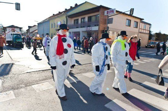 Eine Gruppe von Menschen, die als Schneemänner verkleidet sind, überquert eine Straße bei einem Umzug, mit Zuschauern und Gebäuden im Hintergrund.