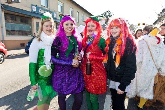 Vier Frauen in bunten Kostümen stehen zusammen auf einer Straße und lächeln in die Kamera. Sie halten Flaschen in den Händen und scheinen sich auf einem Festival zu befinden. Hinter ihnen stehen Gebäude und ein geparktes Auto auf der Straße.