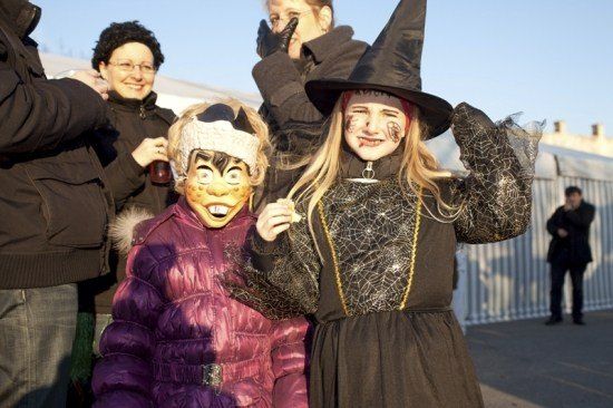 Zwei Kinder in Halloween-Kostümen, eines als Hexe und das andere als Maske, stehen zusammen mit Erwachsenen in einer festlichen Umgebung.
