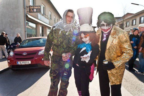Drei Menschen in Kostümen stehen auf einer Straße, einer als Joker, ein anderer als Hutmacher und der dritte in einer Camouflage-Uniform. Ein rotes Auto und ein Gebäude mit einem Schild für Cafe Saletta sind im Hintergrund zu sehen.