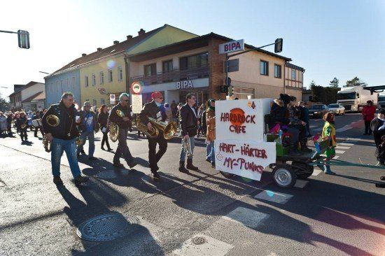 Ein Straßenumzug mit Musikern, die Blechblasinstrumente spielen, und einem Wagen mit einem Schild, das 'Hardrock Cafe' und 'Hart-Harter My-Punks' besagt. Im Hintergrund sind Gebäude und Straßenschilder zu sehen.