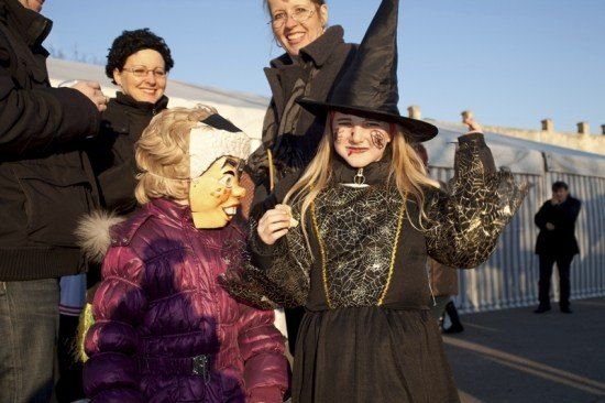 Eine Gruppe von Menschen, darunter zwei Kinder in Kostümen, stehen an einem sonnigen Tag draußen. Ein Kind ist als Hexe und das andere als Hexenmaske verkleidet.