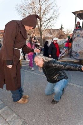 Ein Mann, der als Mönch verkleidet ist, kniet vor einer Frau in einer Lederjacke auf einer Straße während eines festlichen Ereignisses.